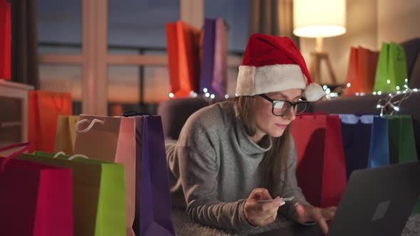 Happy Woman with Glasses Wearing a Santa Claus Hat is Lying on the Carpet and Makes an Online