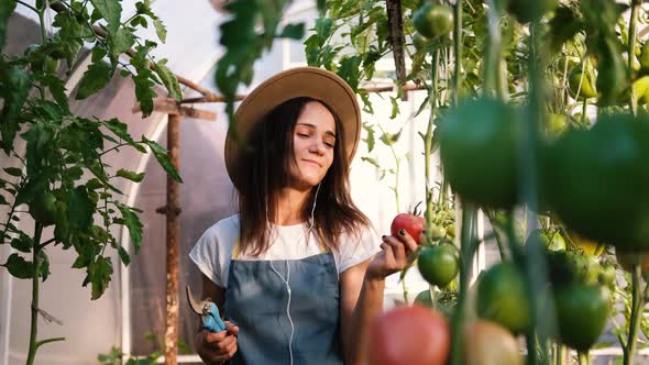 Happy Young Woman Farmer in a Hat Listens to Music on a Smartphone and Sings While Working in a