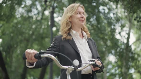 Bottom Angle View of Confident Smiling Businesswoman in Formal Suit Walking with Bicycle in Summer
