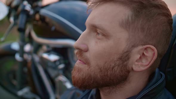 Middleaged Biker with Beard Sits By Motorcycle at Sunset