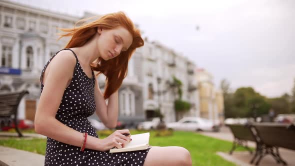 Cute Caucasian Redhead Girl Doing Notes with Hair on Her Face Sitting on Street