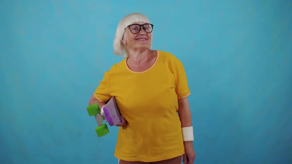 Active Positive Sporty Old Woman in Glasses with Skateboard in Hand on Blue Background