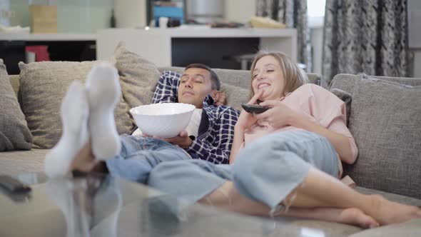 Wide Shot of Relaxed Carefree Couple Lying on Couch Watching Comedy Movie on TV Eating Popcorn