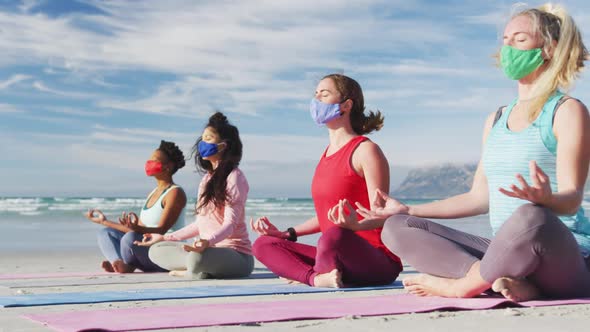 Group of diverse female friends wearing face masks meditating at the beach