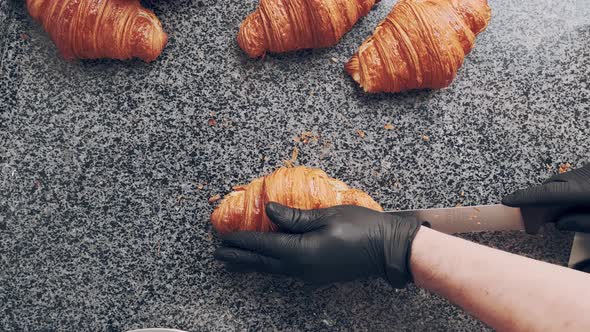 Baker in Black Gloves Cuts Fresh Golden Croissant in Half on Stone Countertop