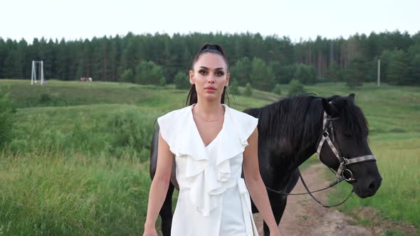 Young Woman Walks and Black Stallion with Bridle Follows