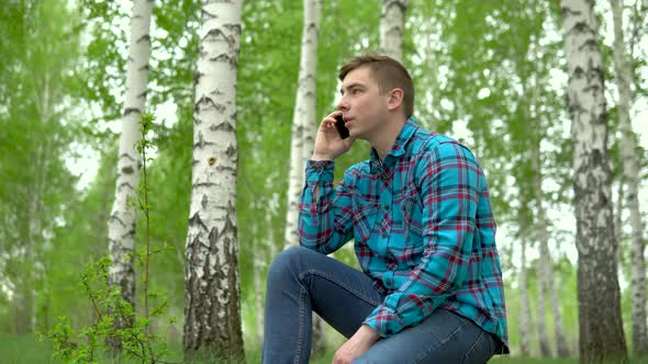 Young Man in Nature with a Phone. A Man Sits on a Stump in a Birch Forest and Talks on the Phone.