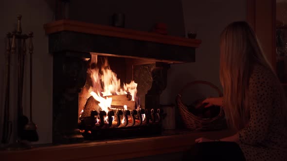 A pretty blonde girl puts a peace of wood into the fire. In the background is a basket with more woo