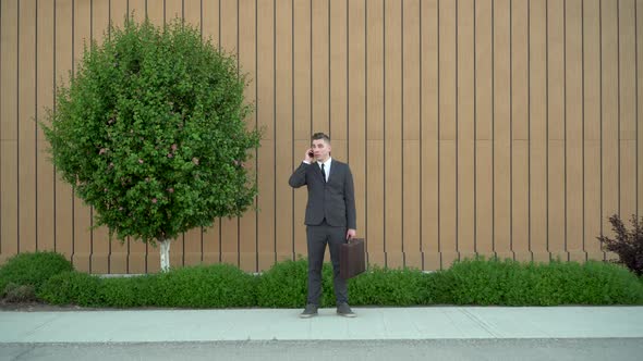 A Young Businessman in a Suit Speaks on the Phone. Serious Man Standing with Briefcase in Hand on