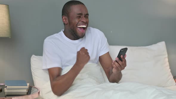 Success, African Man Celebrating on Smartphone in Bed 