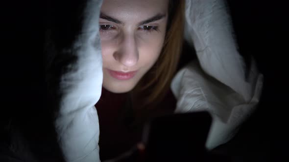 A Young Woman Lies Under a Blanket with a Phone in Her Hands. A Woman in the Dark Uses a Pad. Modern