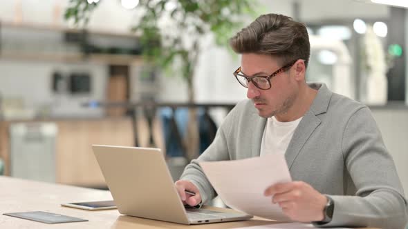 Middle Aged Man with Laptop Reading Documents in Office 