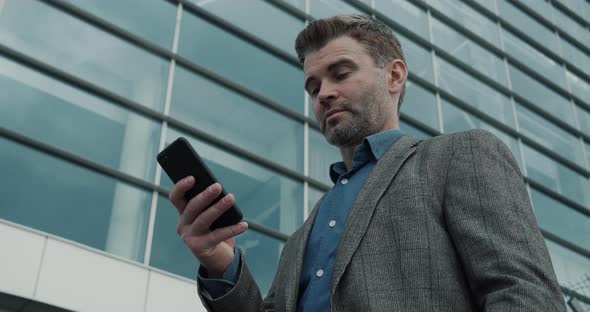 Dreamy confident Businessman working on Smartphone near big modern Office.