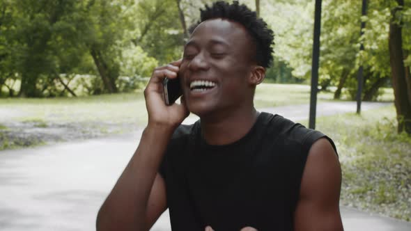 Cheerful Black Millennial Man Talking On Cellphone Sitting Outdoors