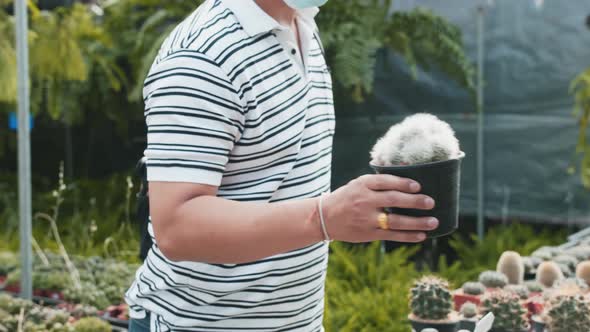 An asian man picks up a cactus in a pot at a plant shop where other people are in the background alo