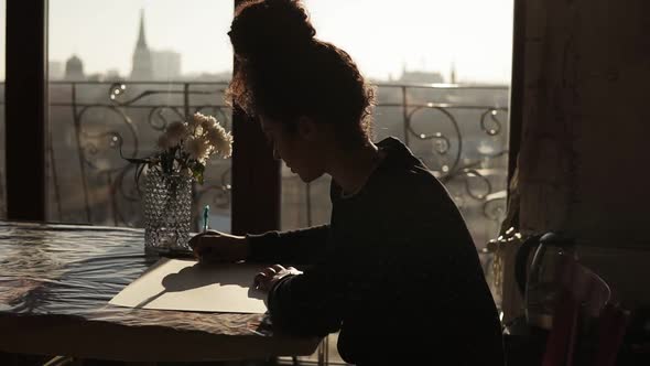 Female Artist is Sitting By the Table with Flowers on It Drawing on Paper with Pen in a Very