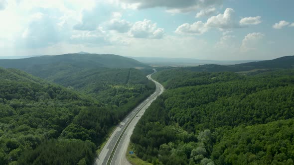 Highway road in green forest hills and blue cloudy sky, aerial pan up