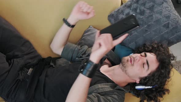 Curly Haired Young Man on the Couch Listening Music with Headphones