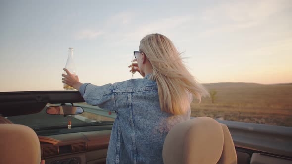 A Young Woman Rides Standing in the Front Seat of a Cabriolet. She Has an Open Champagne Bottle in