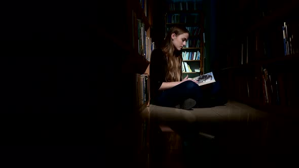 Student Girl Sitting on Floor and Examines Book in Library, Camera Moves Left To Right