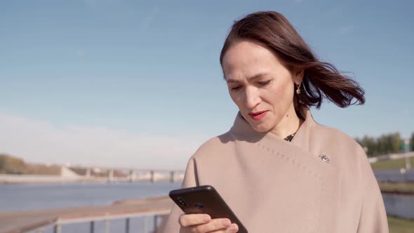 A Business Lady in a Coat Looks Through Messages on a Mobile Phone on the Street