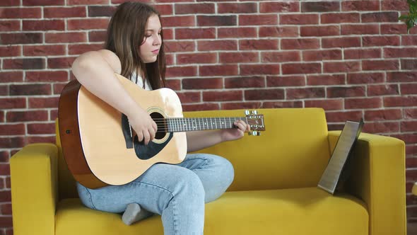 Young Woman is Sitting on Sofa and Playing Acoustic Guitar According to Notes