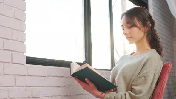 Beautiful Woman Sitting Near Window and Reading a Book on Background of Sunrise