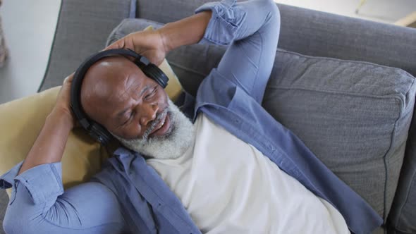 Relaxed senior african american man in living room lying on sofa, wearing headphones