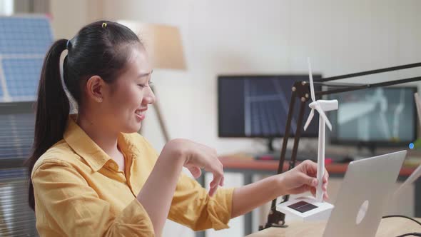 Asian Woman Sitting In Front Of Solar Cell Looks At Wind Turbine While Typing On A Laptop