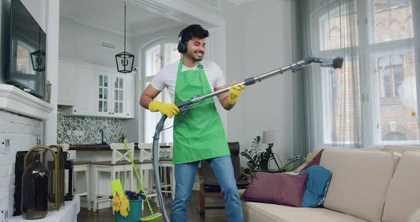 Young Cleaner in Uniform which Listening Music in Headphones while Vacuuming Sofa Dust Mites