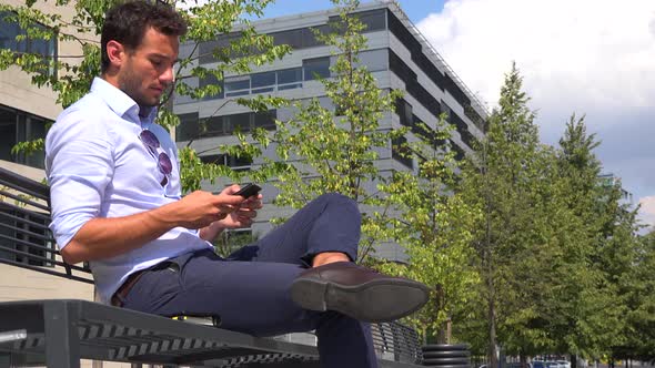 A Young Businessman Sits on a Bench and Works on a Smartphone, View From Below, an Office Building