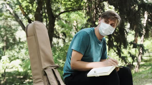 Young Man Is Sitting on a Bench in Park and Write Some Song Ideas in Notebook