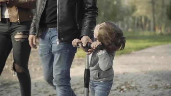 Cheerful Cute Caucasian Boy Riding Scooter in Sunny Park with Unrecognizable Parents