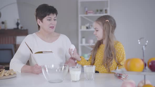 Portrait of Diligent Little Girl Helping Grandmother with Baking of Pancakes for Shrove Tuesday