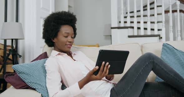 Woman with African Hairstyle Lying on Couch and Enjoying from Review of Apps on Tablet Device