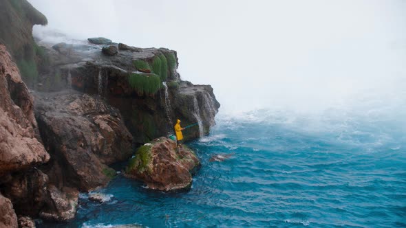 A Man in a Yellow Raincoat Fishing in the Blue Sea Standing By the Unrestrained Waterfall