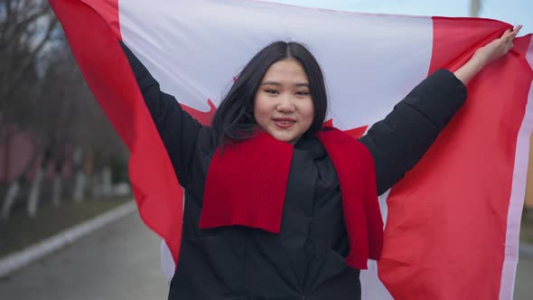 Cheerful Asian Woman Holding Canadian Flag in Stretched Hands Looking at Camera Smiling and Wrapping