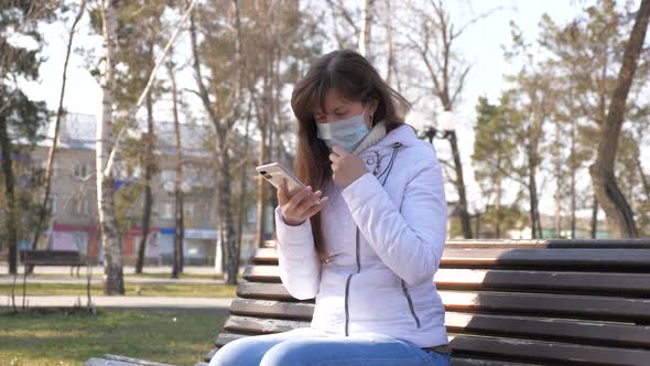 Young Woman Traveler in Medical Protective Mask Sits with Smartphone on City Street