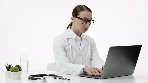 Female Caucasian Doctor in White Medical Coat, Glasses Works in Office at Laptop