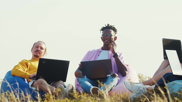 Caucasian and AfricanAmerican Man Sitting on Chairs Bags with Laptops on Their Knees in Nature Front