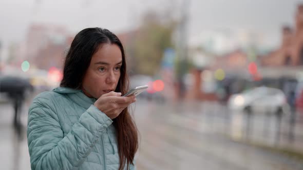 a Brunette in a Blue Jacket and with Long Hair Stands and Says a Message Into Phone on a Wet Street