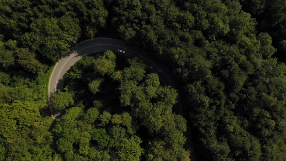 Aerial view at a white car from above drining into a tight curve in a forest, look up shot to a curv