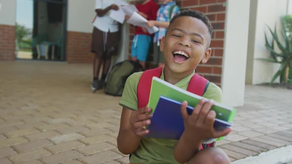 Video of happy african american boy holding books in front of school