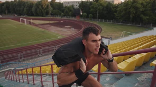 A Naked Torso Bodybuilder Training in City Stadium and Doing Squats with a Sandbag on His Shoulders