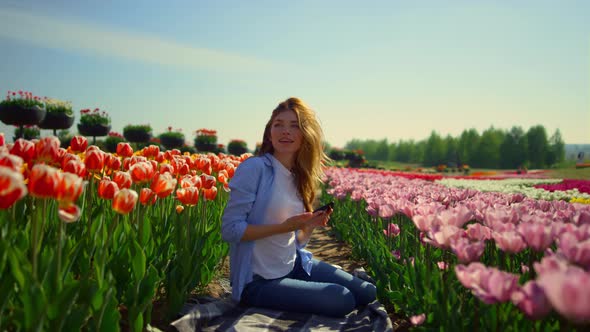 Young Woman Sitting in Blooming Flower Garden with Mobile Phone in Sunny Day