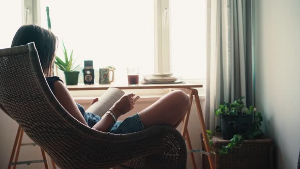 A Woman Reading a Book While Sitting in a Comfortable Wicker Chair