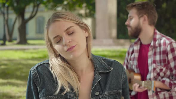 Close-up Face of Young Blond Woman Listening To Music and Smiling. Blurred Romantic Man Playing