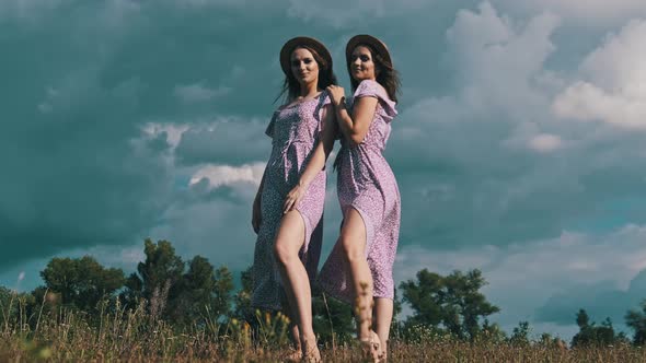 Two Young Slender Twin Girls in Identical Dresses Looking at Camera in Nature