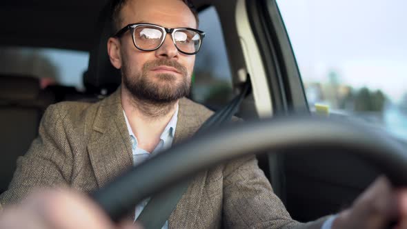 Satisfied Bearded Man in Glasses Driving a Car Down the Street in Sunny Weather