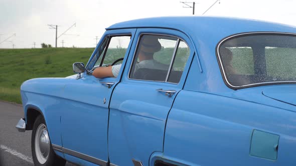 Young Couple in Hats Driving at Blue Retro Car After Rain During Summer Travel. Unrecognizable Pair